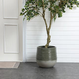 Tall potted plant in a textured gray planter with a drainage hole, on a dark tiled floor, placed against a white paneled wall next to a door with molding.