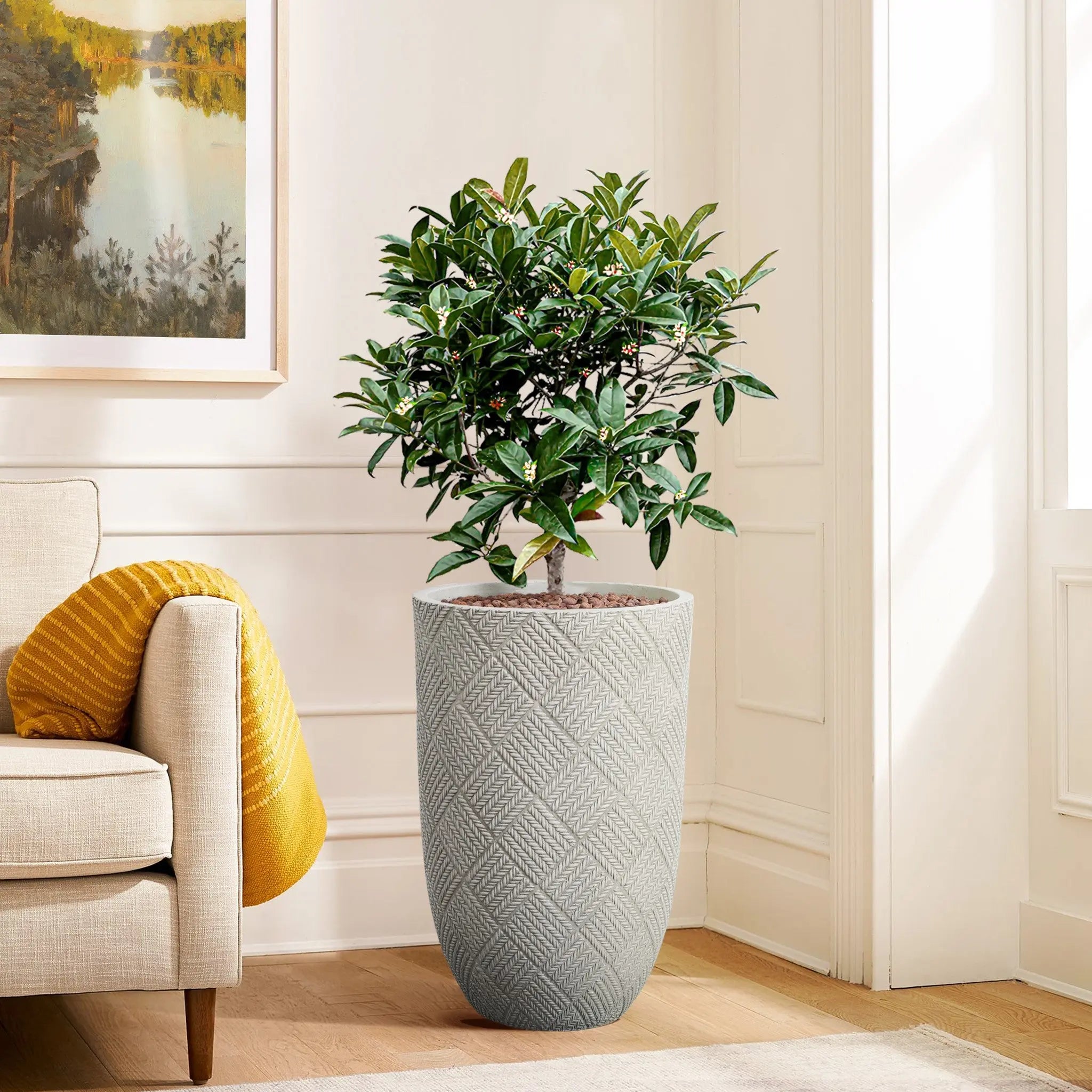 A potted plant in a textured gray planter, boasting modern minimalist design, sits next to a beige sofa with a yellow pillow, while a framed landscape painting adorns the wall.