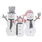 Three snowmen with black and white scarves, smiling faces, and red accessories stand together as a charming outdoor Christmas decor; one holds a small wrapped gift.
