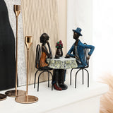 Two modern brown resin sculptures of a couple sitting at a table with a flower vase, creating romantic moments, are displayed on a shelf next to tall candle holders.