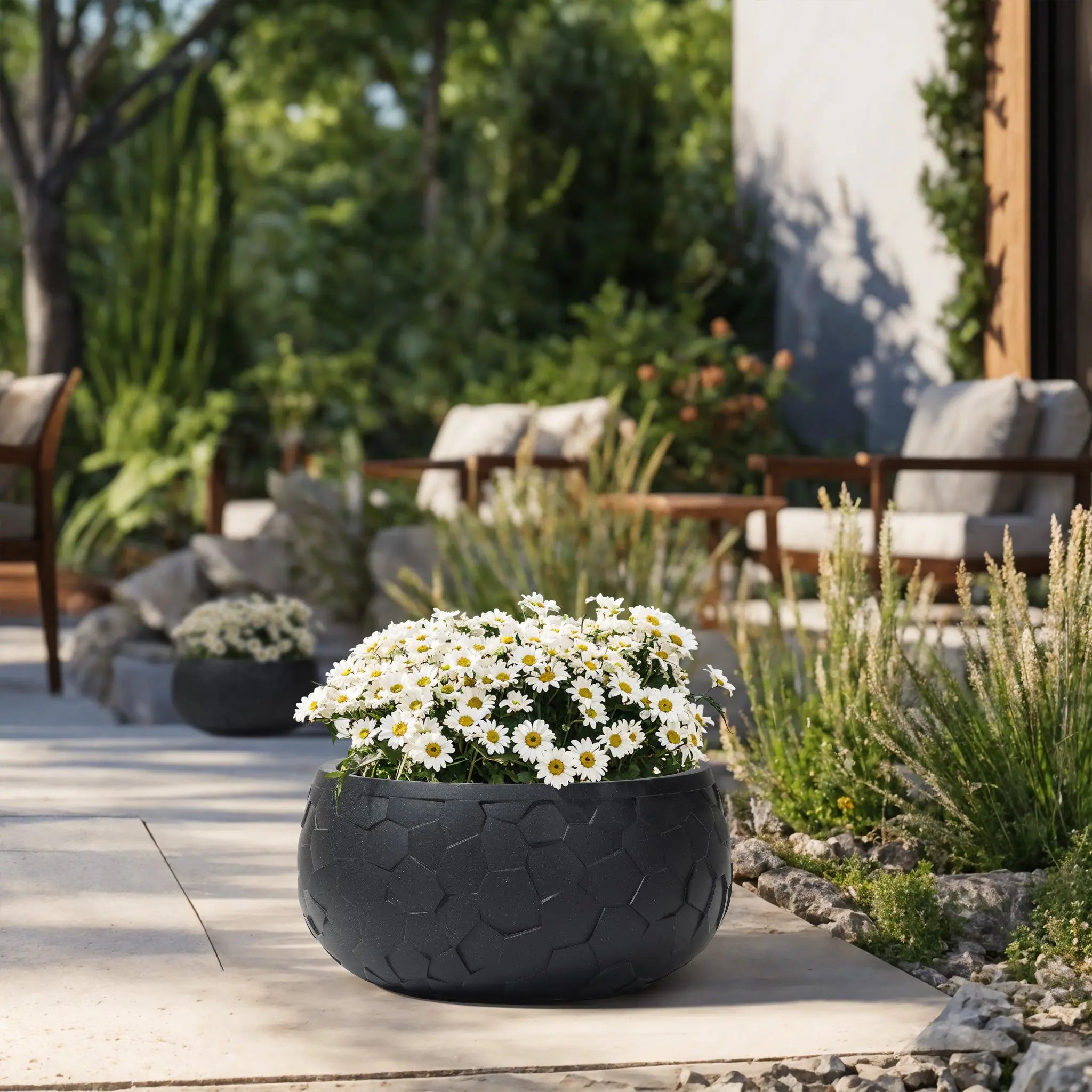 A large LuxenHome Black beehive pattern round tabletop planter, 17.5 wide, with white daisies sits on a sunlit patio. Wooden chairs and lush green plants create a serene outdoor scene in the background.