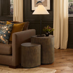 A living room scene exuding rustic charm features teardrop accent tables, a table lamp, and a small vase with green leaves, all elegantly placed next to a brown sofa adorned with patterned and solid pillows.