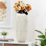 A LuxenHome 17 Modern geometric pattern resin tall vase, filled with orange and cream flowers, sits on a white tabletop beside a plant. In the background, a framed abstract art piece with an antique effect is displayed alongside a small stack of books.