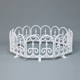 Small white circular fence with decor scroll ,pointed designs, against plain gray background.