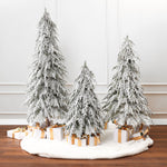 3 snow-flocked cedar trees are arranged on white circular rug with wrapped gifts in white ,gold underneath, againstwhite paneled wall.