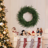 Artificial wreath hangs on a white wall above a mantel adorned with stockings, nutcrackers, plush toys, lit lantern.