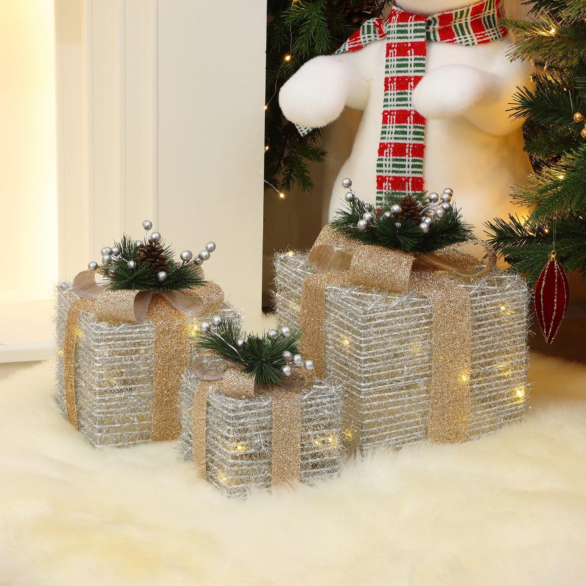 3 silver tinsel gift boxes with bows, pine accents are arranged under Christmas tree on white furry rug.