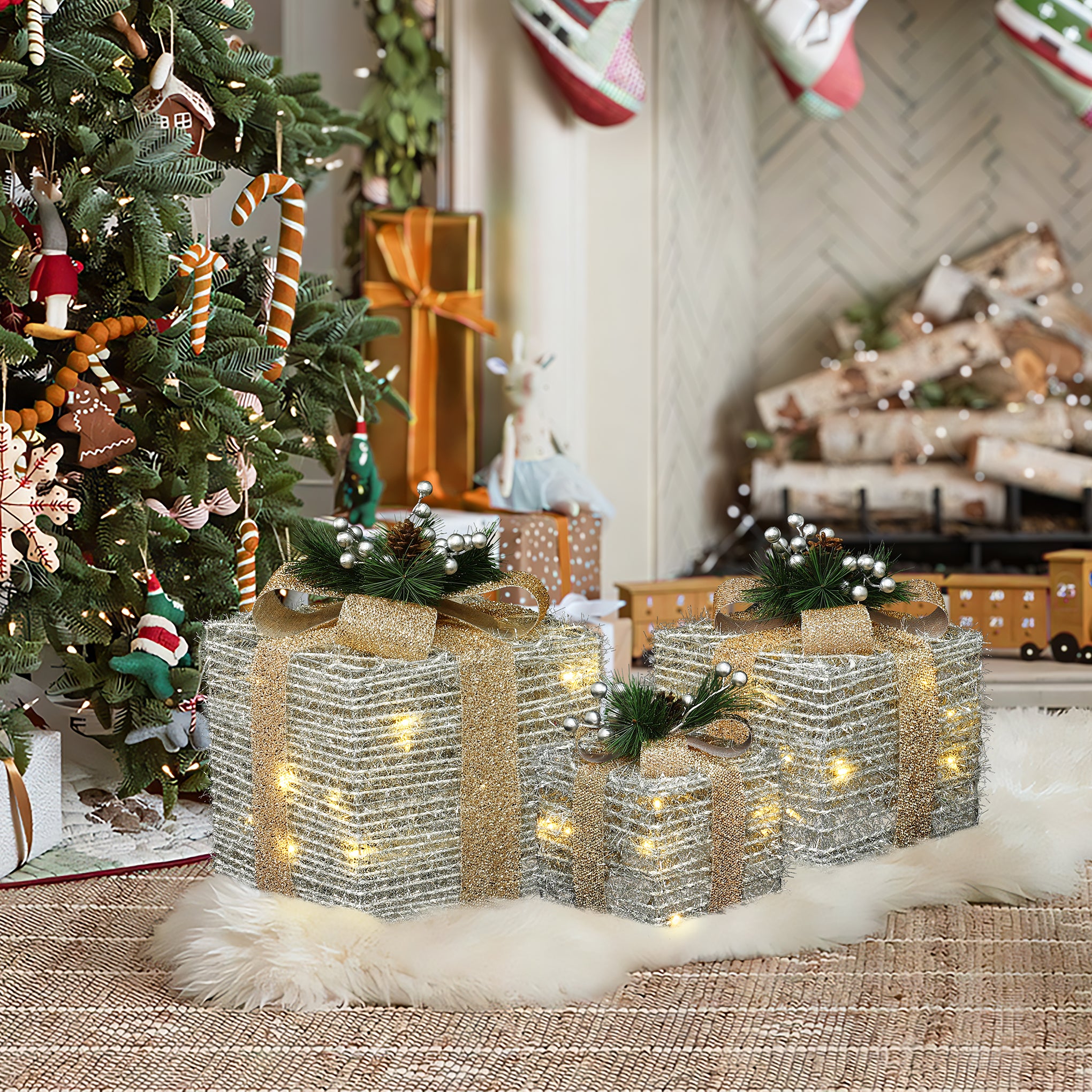 3 silver tinsel gift boxes,gold ribbons,lights sit on white rug near christmas tree, fireplace adorned with stockings.