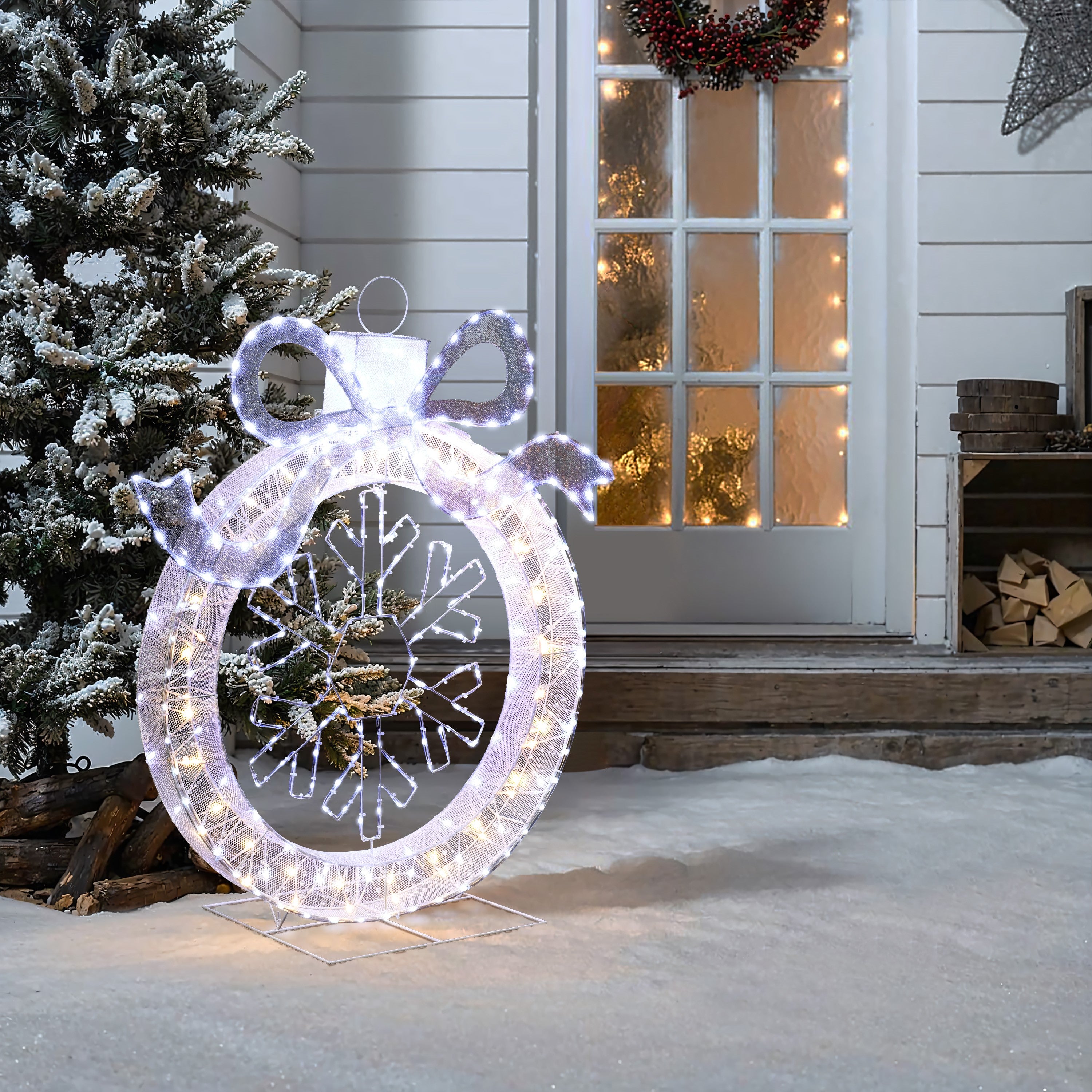 Large lighted ornament with snowflake stands on snowy ground, near holiday decor,lit christmas tree with wreath.