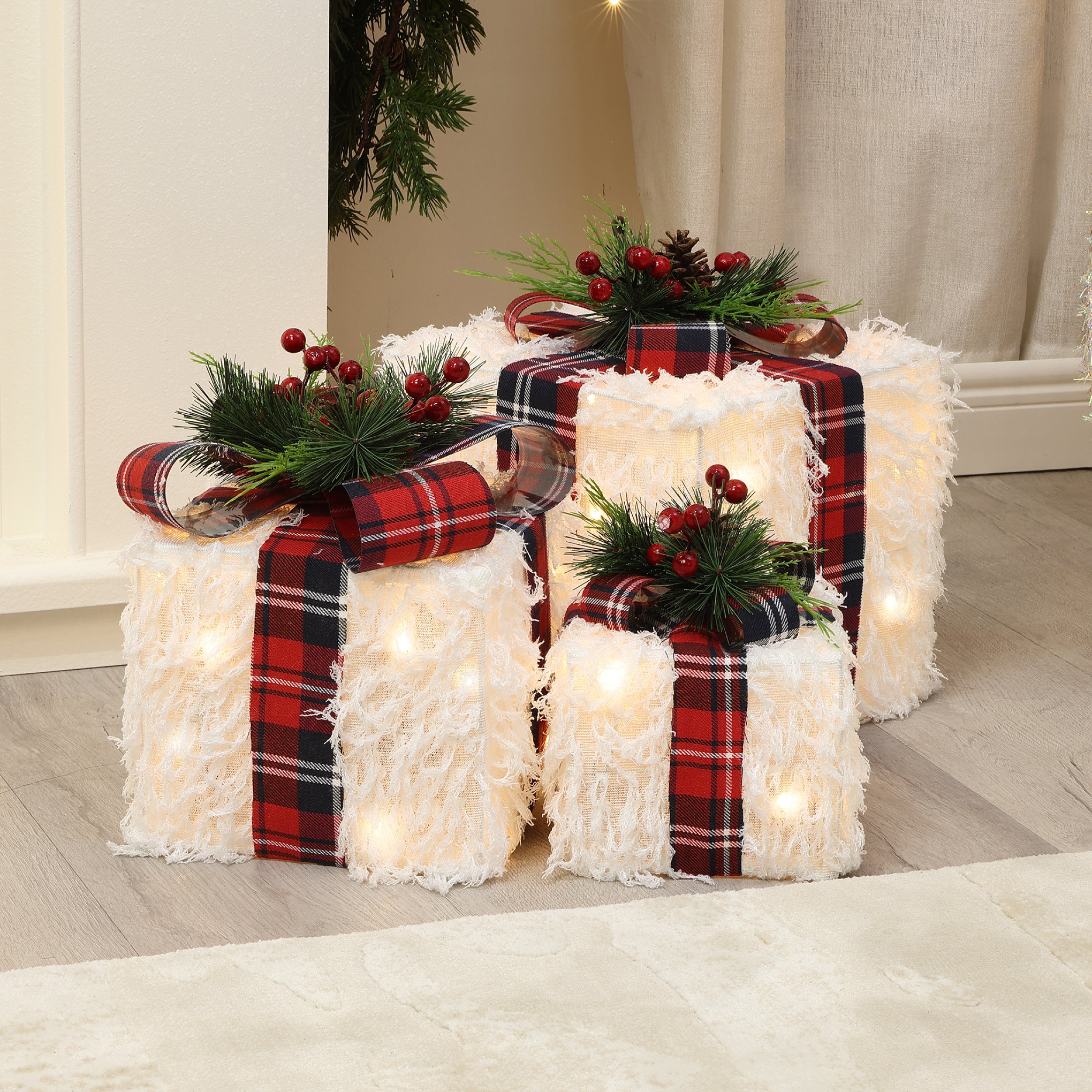 3 gift box decor pieces with white textured, plaid ribbons, greenery, red berries on wooden floor near wall.