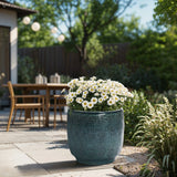 An 11.22 high Marina gray glazed round ceramic planter by LuxenHome, filled with blooming white daisies, graces the patio. In the background, a wooden dining set, lush greenery, and hanging string lights enrich the serene garden ambiance beneath a gradient-colored sky.