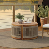 Round acacia wood patio table with hand-woven rope detailing, adorned with a potted plant, stack of books, and candle holder, set on a circular rug. Versatile for both indoor and outdoor settings, the scene is completed by a railing and lush foliage in the background.