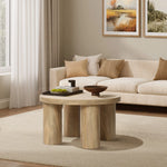 A cozy living room with a beige sofa, oak-finish coffee table, decorative bowl, books, and a green vase. Two landscape paintings hang on the wall above the sofa.