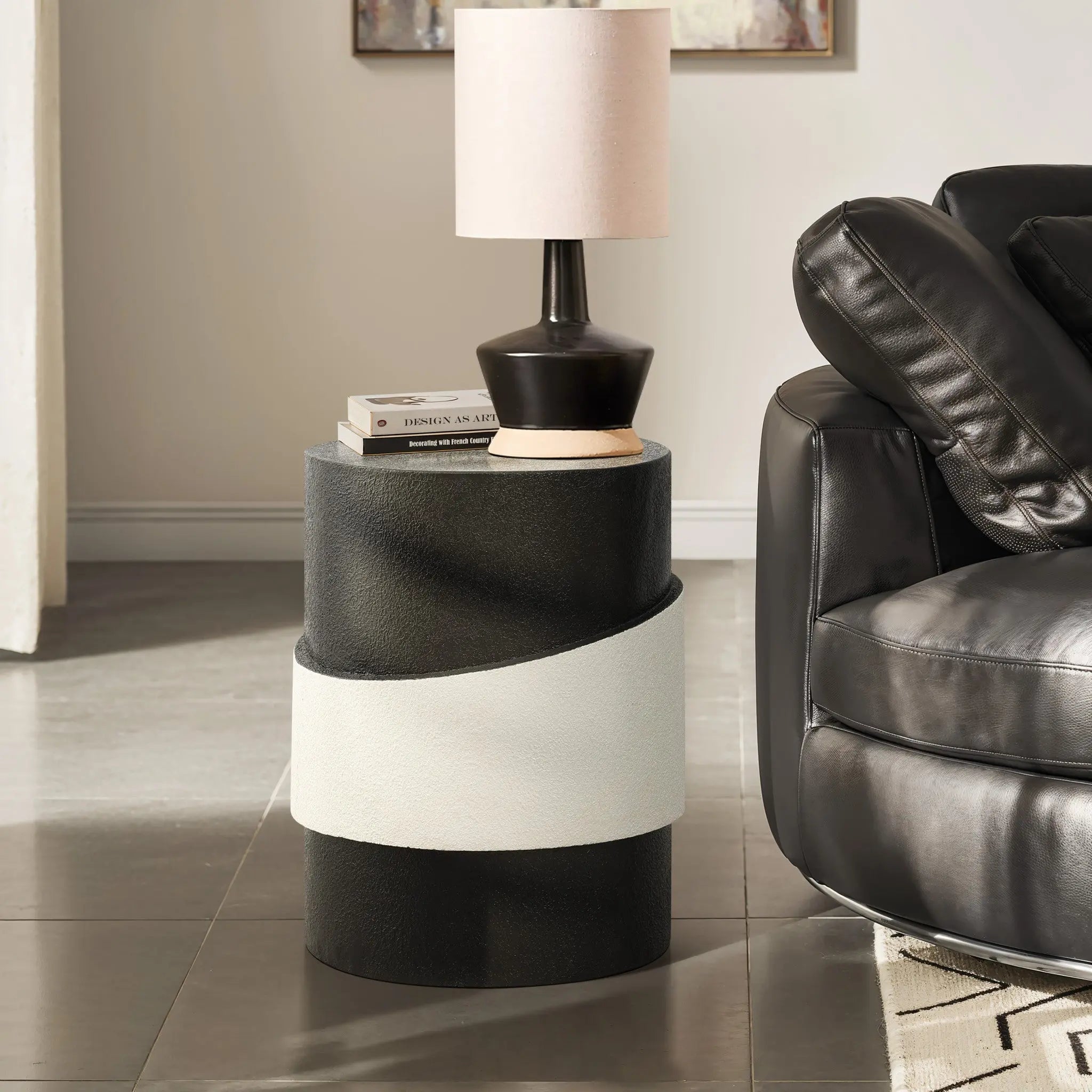 A small round side table with a unique white irregular pattern holds a lamp and books, perfectly positioned next to a black leather sofa in the tiled room.