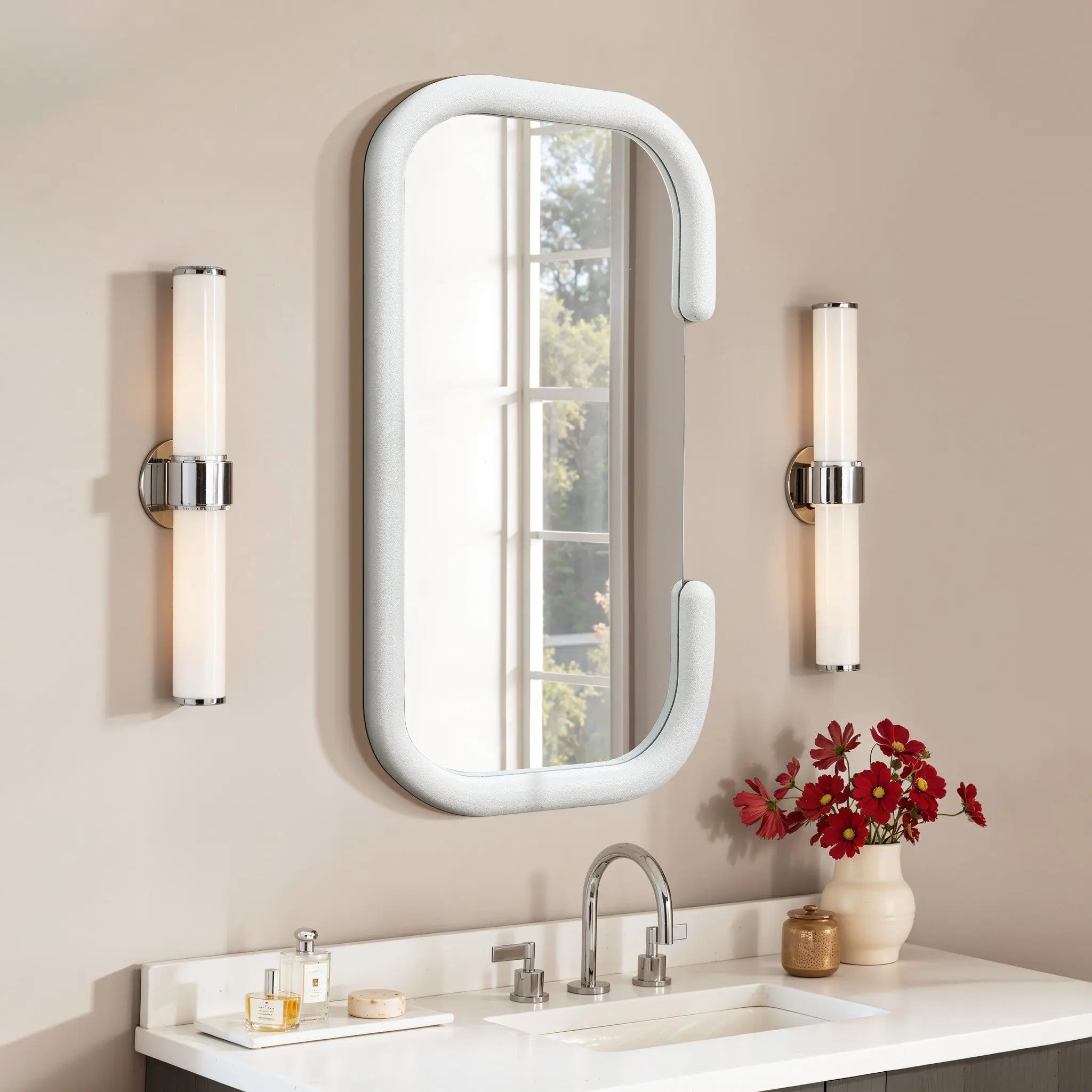Bathroom vanity with a contemporary C-shaped silhouette mirror, two vertical light fixtures, and a sink. A vase with red flowers is placed on the counter alongside toiletries, bringing modernist elegance to the space.