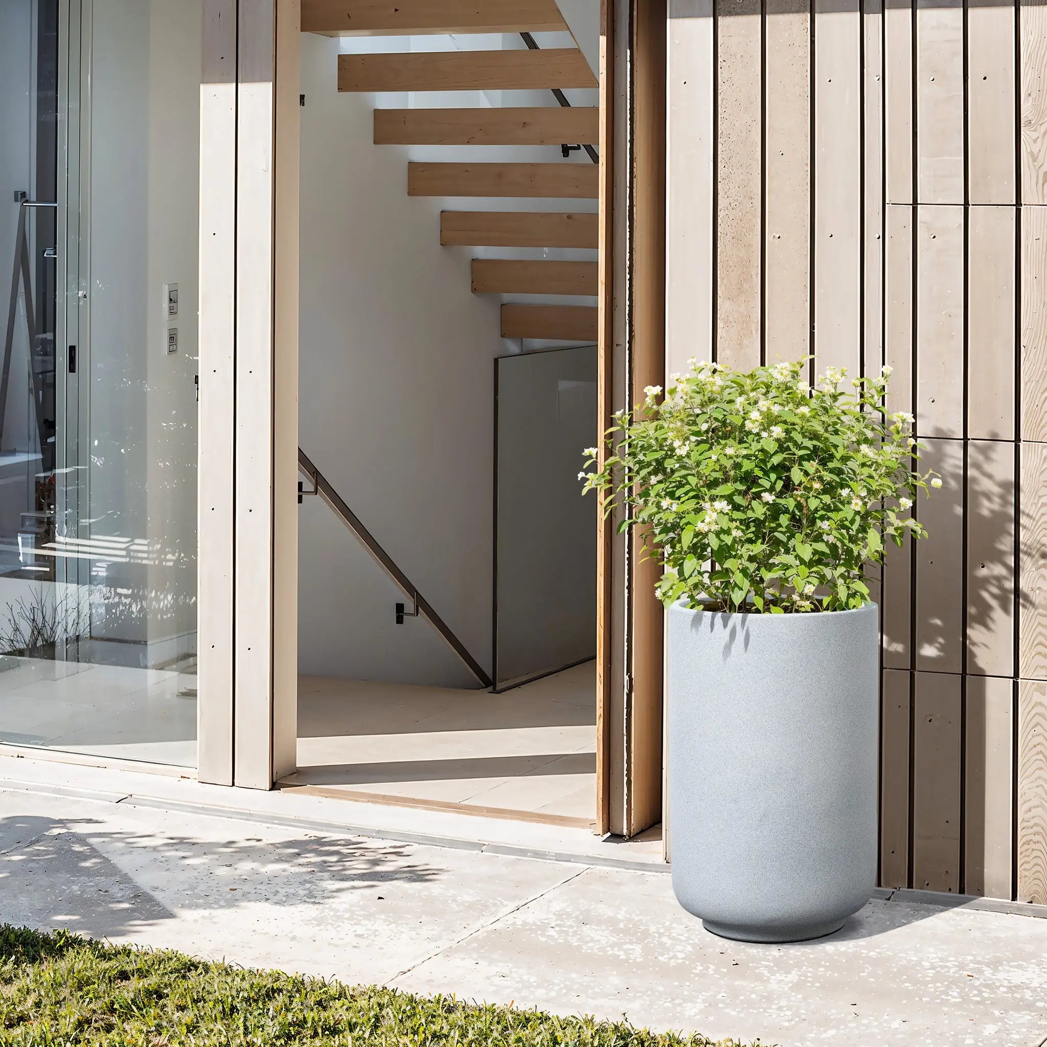 A modern outdoor space features a weather-resistant Gray Round Planter holding a vibrant green plant. Behind, sleek glass doors open to reveal wooden stairs ascending, embodying the essence of modern minimalist decor.