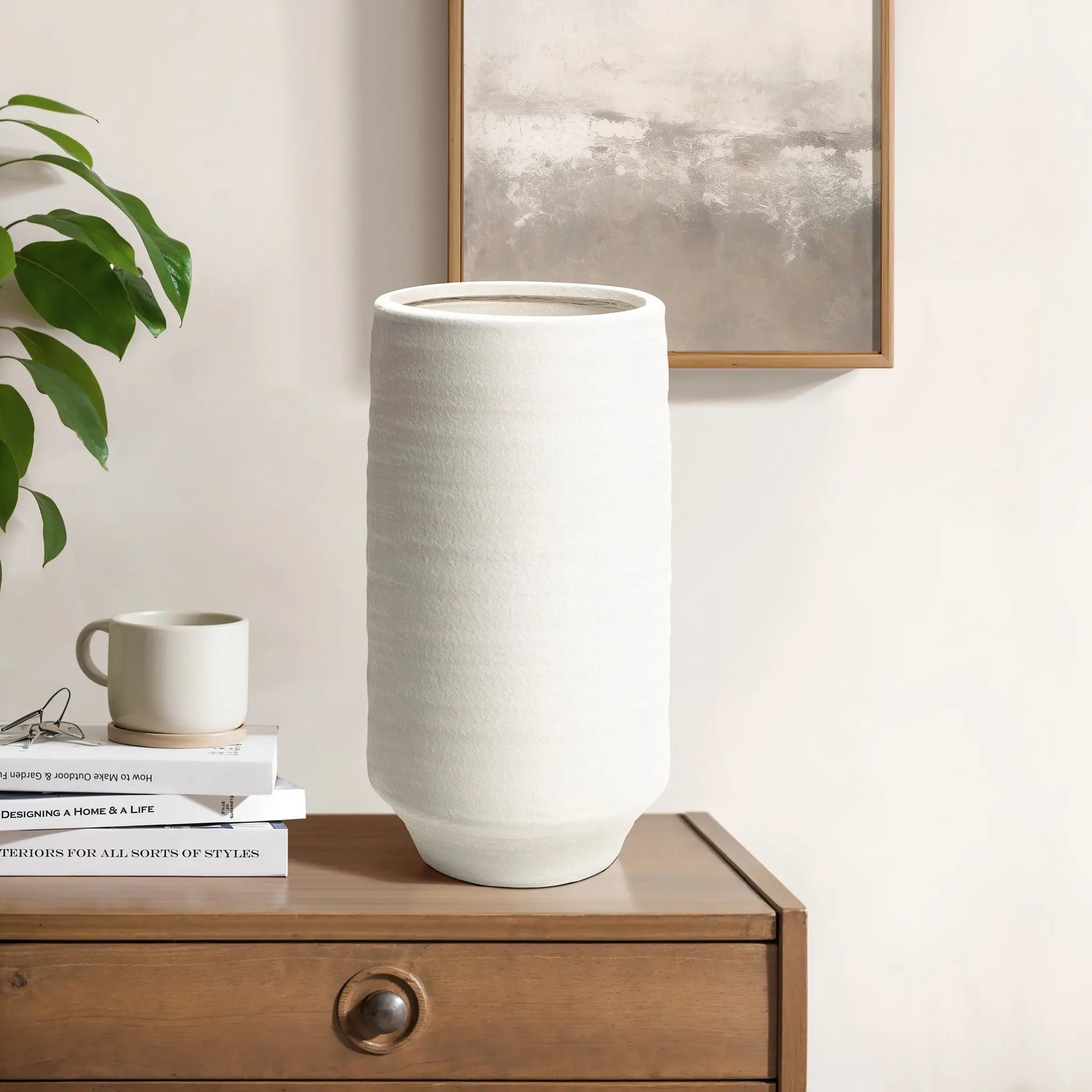 A tall, white terracotta vase graces a wooden dresser, surrounded by books and a mug. A plant sits nearby, with a framed abstract painting hanging on the wall in the background.