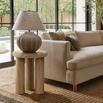 A beige couch is next to a round side table crafted from manufactured wood, featuring an oak finish. The setting includes a woven rug and large windows with a garden view, completed by a textured lamp on the table.