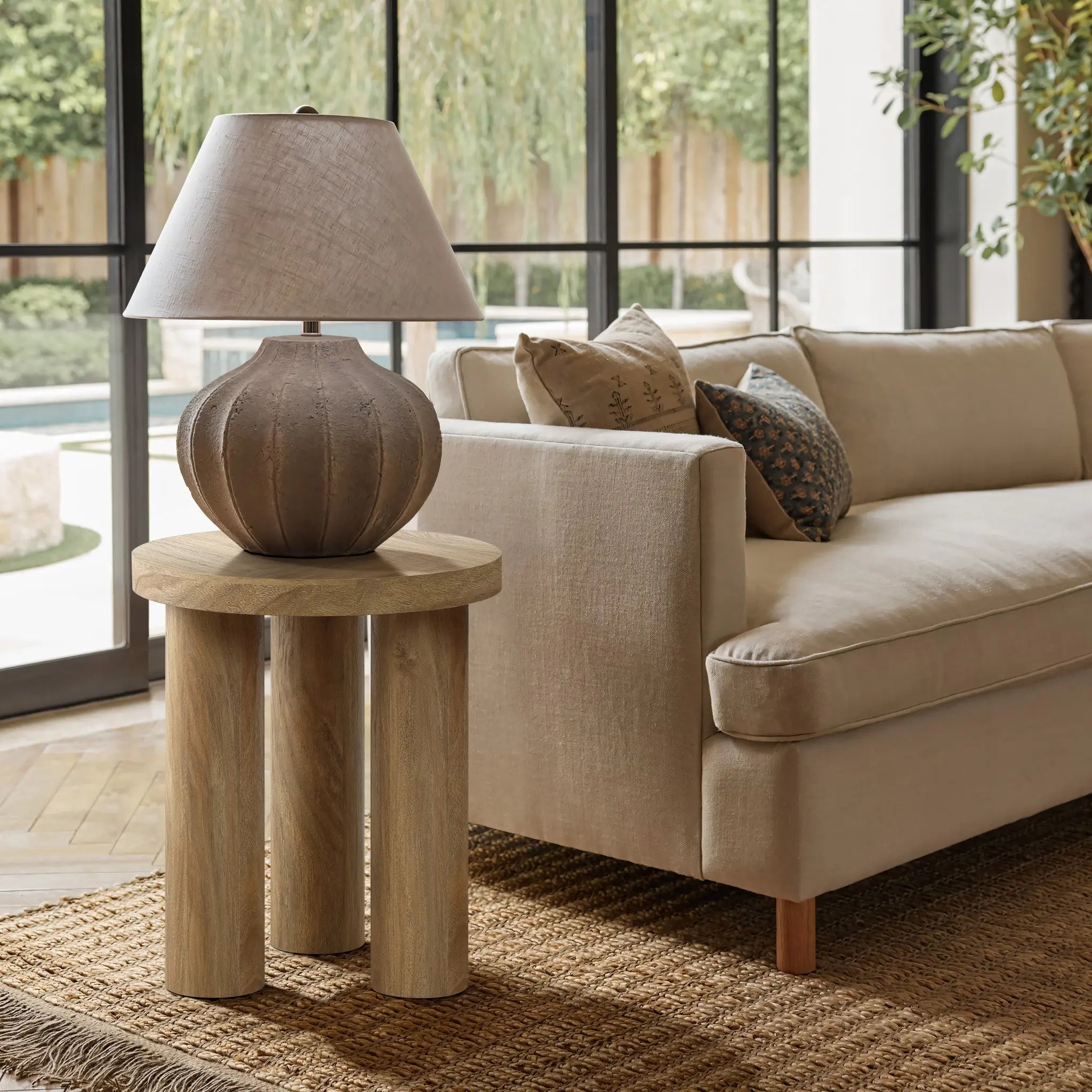 A beige couch is next to a round side table crafted from manufactured wood, featuring an oak finish. The setting includes a woven rug and large windows with a garden view, completed by a textured lamp on the table.
