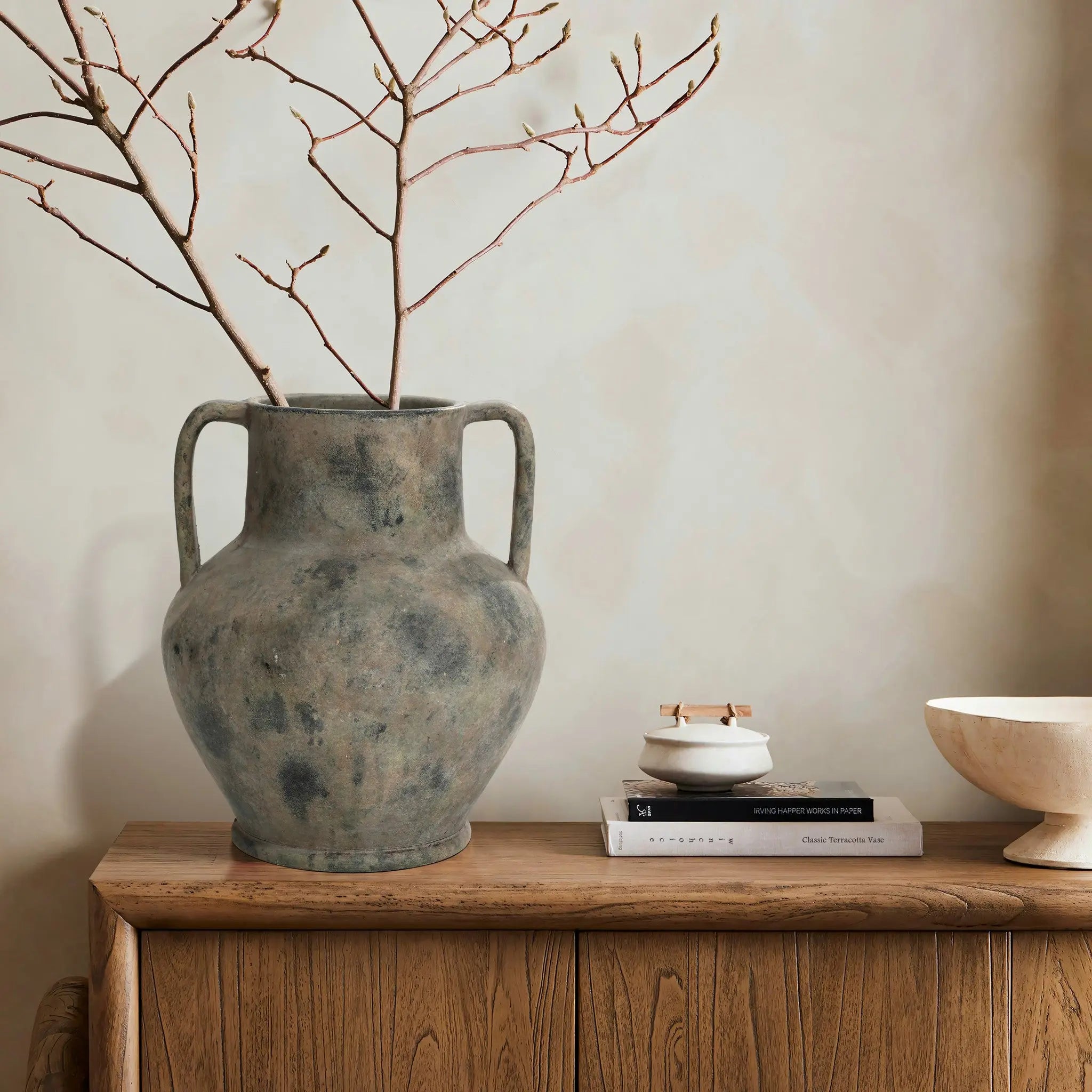 A large LuxenHome rustic mottled grey terracotta urn vase with handles, 12.1 h, holds bare branches atop a wooden cabinet. Beside it are a stack of books, a small lidded jar, and an off-white bowl, set against a neutral wall.