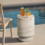A wooden chair is beside a round stone table by the pool, embracing both indoor and outdoor aesthetics. The table, reminiscent of a travertine-style side table, holds a tray with a pitcher of iced tea, a glass, and a small potted plant.
