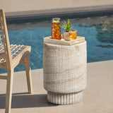 A wooden chair is beside a round stone table by the pool, embracing both indoor and outdoor aesthetics. The table, reminiscent of a travertine-style side table, holds a tray with a pitcher of iced tea, a glass, and a small potted plant.