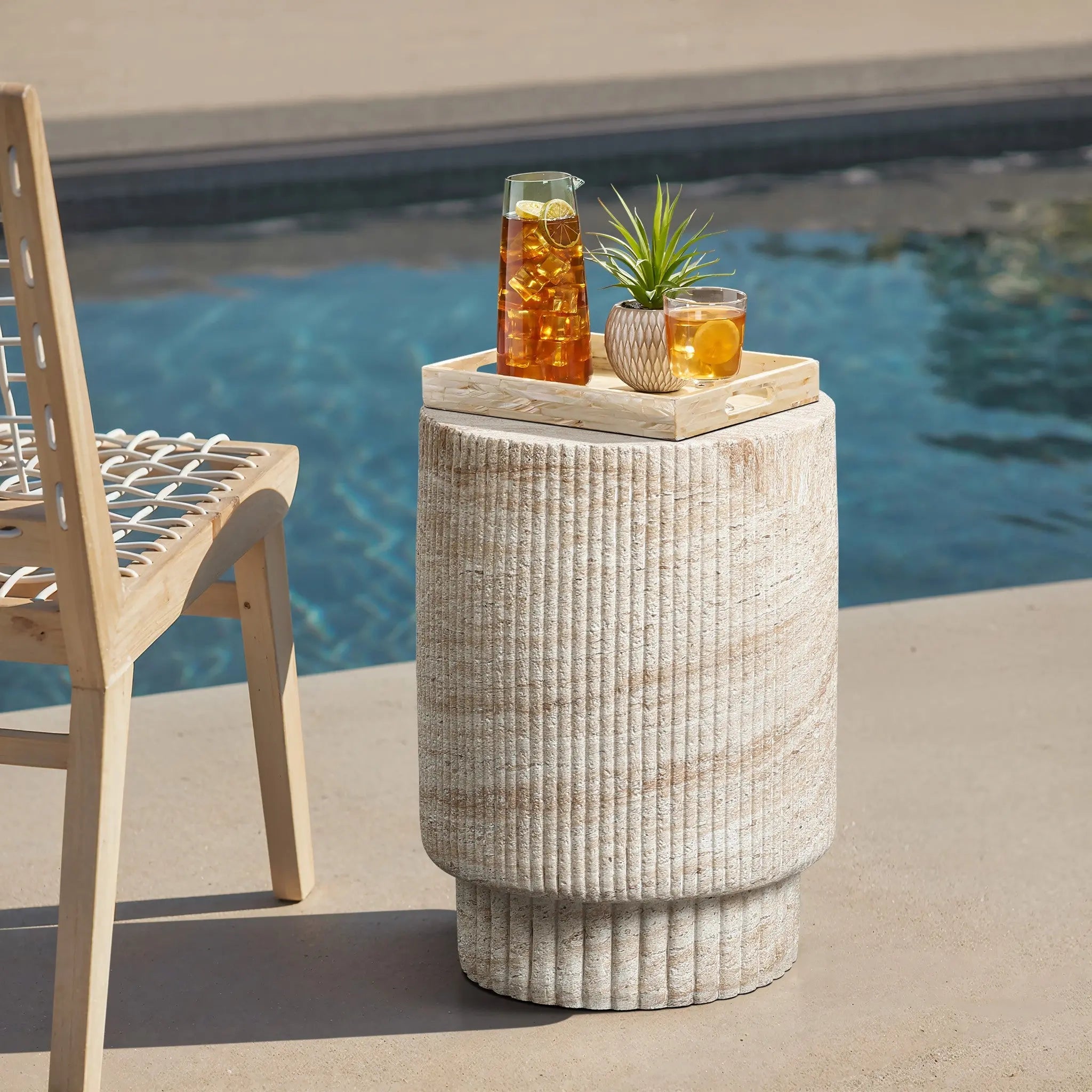 A wooden chair is beside a round stone table by the pool, embracing both indoor and outdoor aesthetics. The table, reminiscent of a travertine-style side table, holds a tray with a pitcher of iced tea, a glass, and a small potted plant.