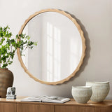 A round mirror with a scalloped design and wooden frame hangs above a wooden console. A potted plant, ceramic bowls, and an open book rest on the console as shadows from a window dance across the wall.