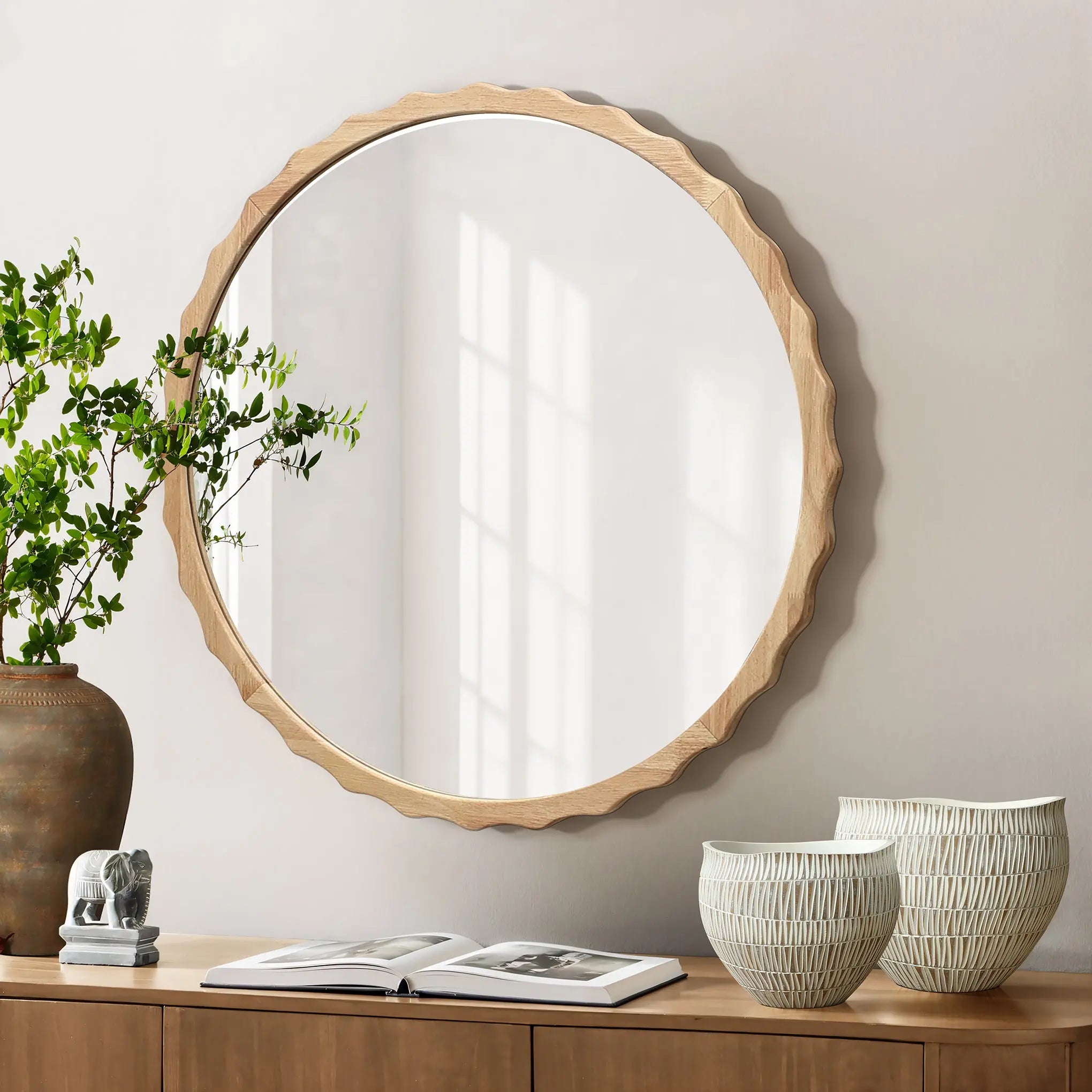 A round mirror with a scalloped design and wooden frame hangs above a wooden console. A potted plant, ceramic bowls, and an open book rest on the console as shadows from a window dance across the wall.