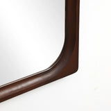 Close-up of a corner of a rectangular wall mirror with a dark brown rubberwood frame, enhancing the home decor against a white wall.