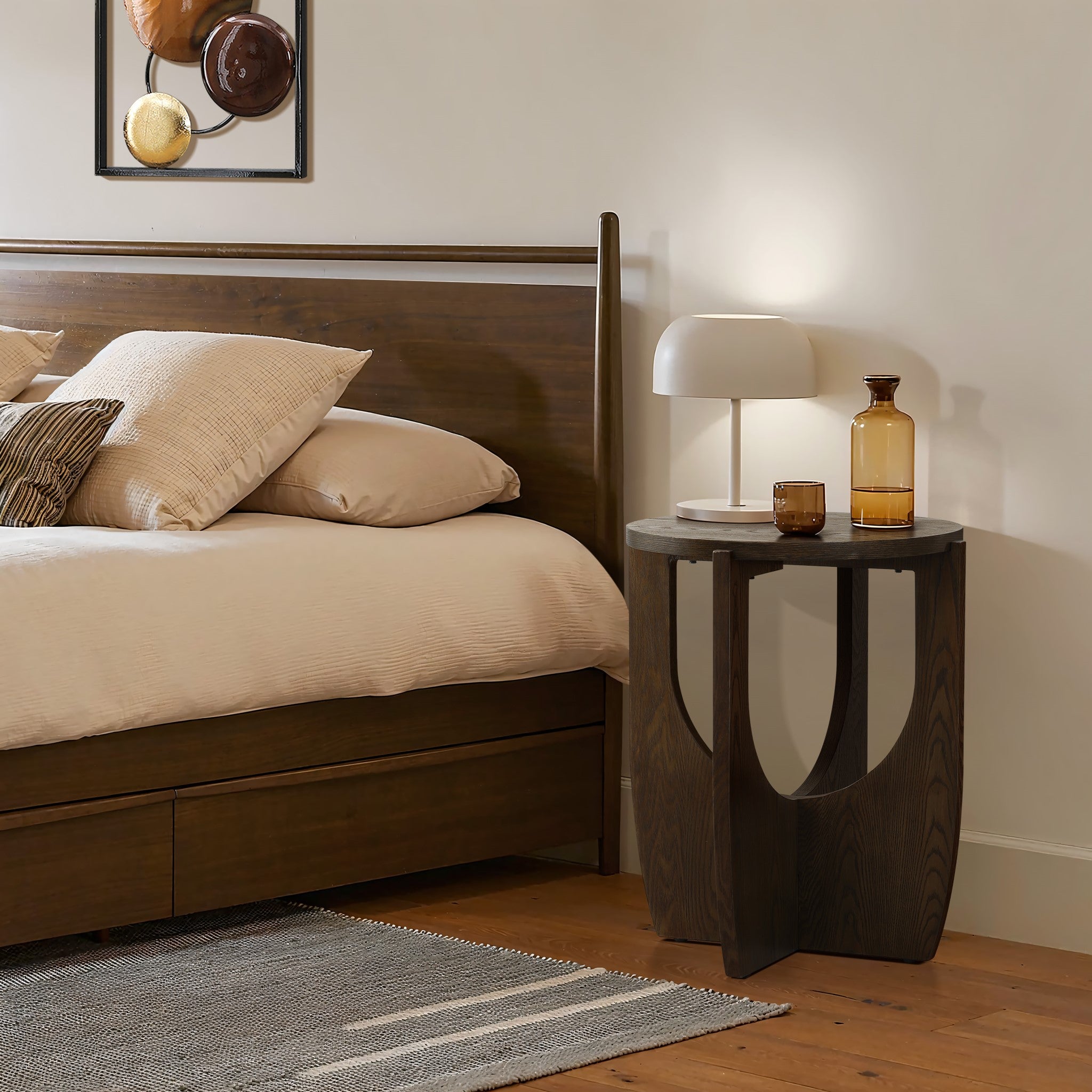 A bedroom scene featuring a wooden bed with beige bedding and a mid-century modern side table. The side table, adorned with two glass bottles and a white lamp, complements the space beautifully. Above the bed hangs a framed abstract artwork, adding an artistic touch to the serene setting.