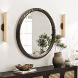 An Aged wood finish crater pattern round wall mirror by LuxenHome is mounted on a white wall between two cylindrical sconces. Below, a sideboard holds a potted plant, a tray with small balls, and a sculpted item, reflecting an industrial-inspired design through the bright window.