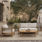 Outdoor patio with beige cushioned chairs, a Rope Sides Loveseat, wooden coffee table with teapot and cups, potted plants, and an umbrella on a woven rug—Club-Style Patio Furniture set against stucco walls and trees.