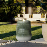 A lit outdoor gas lamp with a ribbed design stands on a patio with a grassy area and wicker seating in the background, subtly illuminated by LED lights.