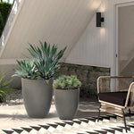 A modern planter set featuring two large potted plants graces the patio, complemented by a wicker chair against the white house backdrop.
