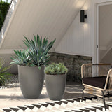 A modern planter set featuring two large potted plants graces the patio, complemented by a wicker chair against the white house backdrop.