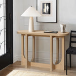A modern living room corner features the LuxenHome Lorean natural finish contemporary console table with a unique curved design. It holds a ceramic lamp with a pleated shade, a decorative cup, and a small book. An abstract art piece is beautifully framed above it.