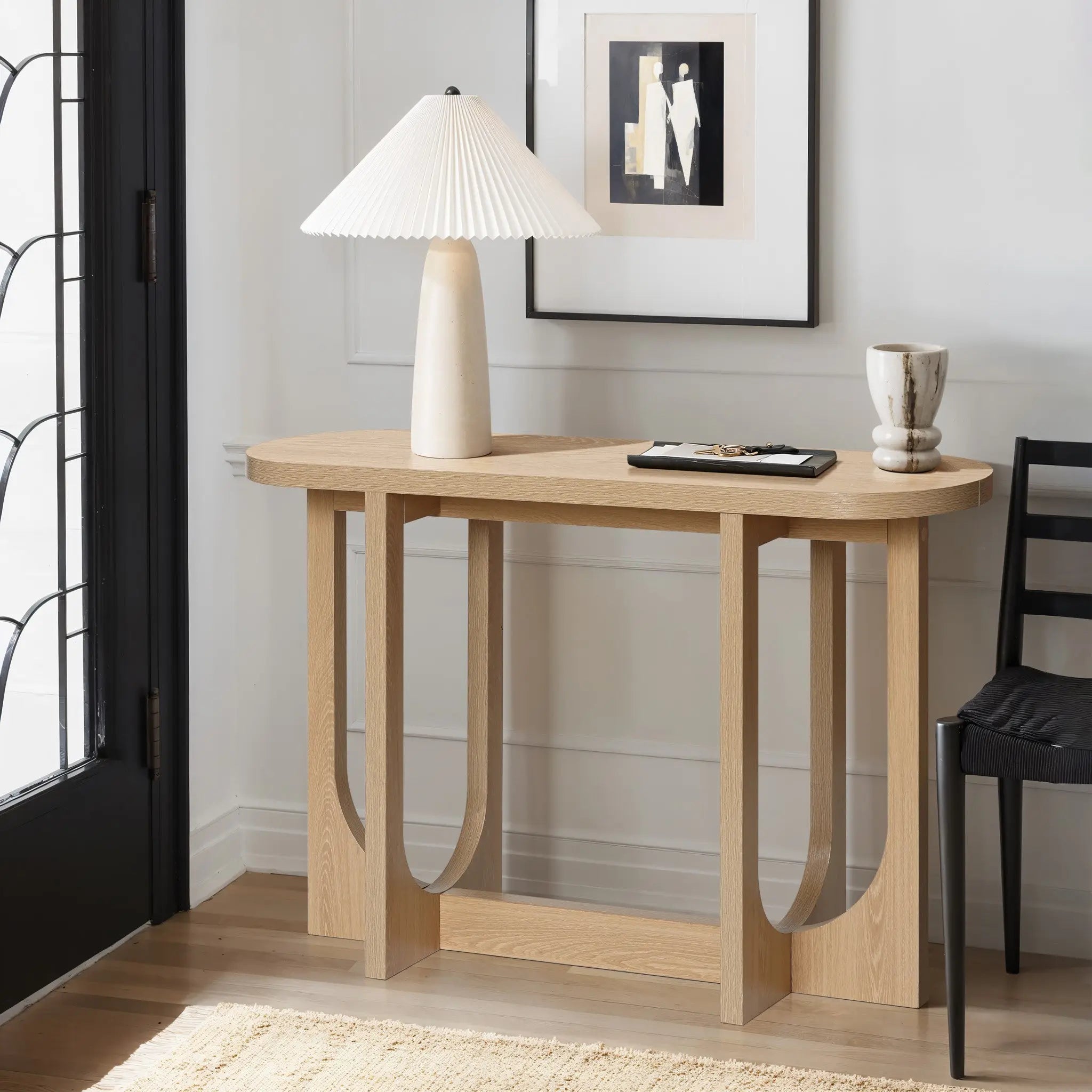 A modern living room corner features the LuxenHome Lorean natural finish contemporary console table with a unique curved design. It holds a ceramic lamp with a pleated shade, a decorative cup, and a small book. An abstract art piece is beautifully framed above it.