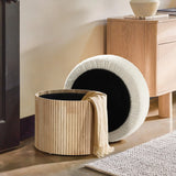 The Mina boucle and wood storage ottoman by LuxenHome features a stylish removable lid, showcasing its storage function when the light brown throw is removed. In the background, an elegant touch is maintained with a small wooden cabinet and a vase on the smooth concrete floor.