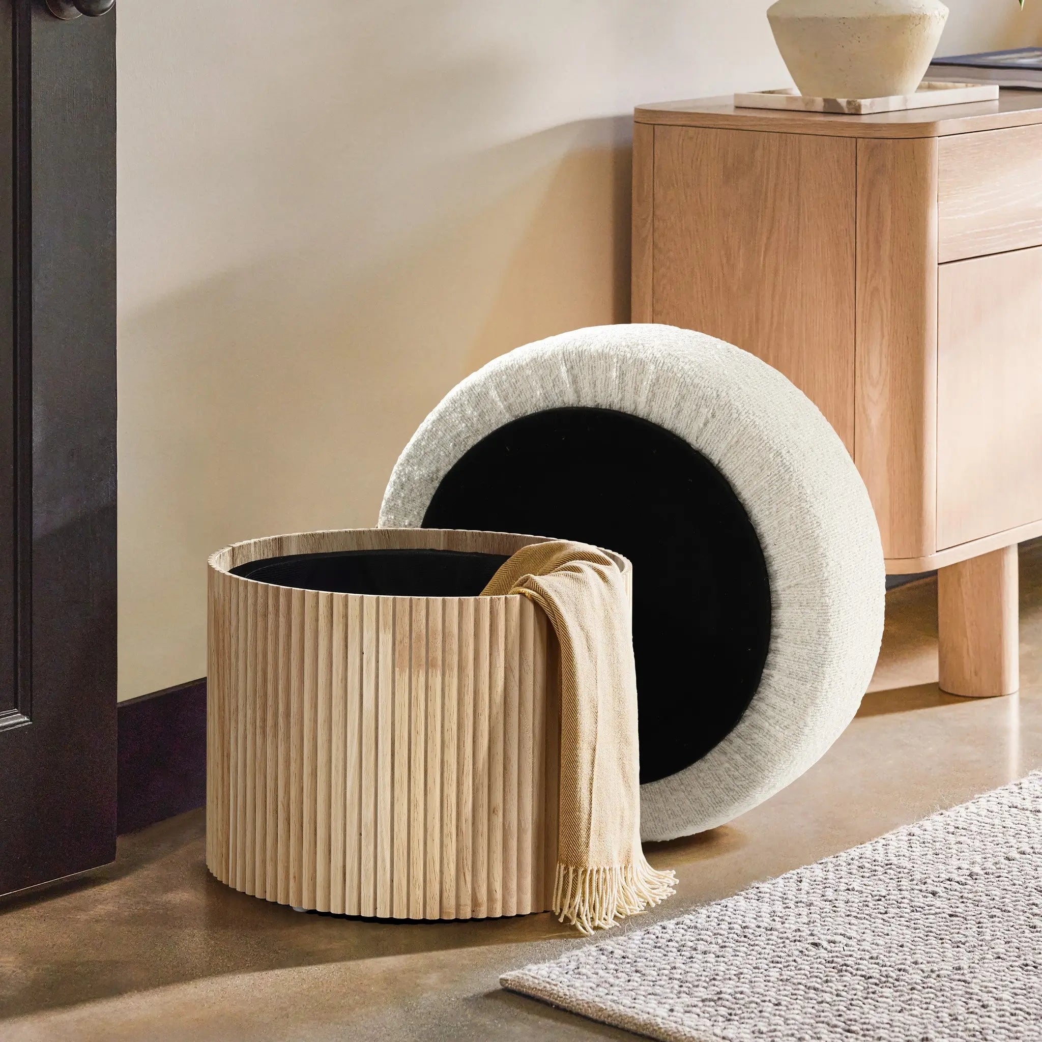 The Mina boucle and wood storage ottoman by LuxenHome features a stylish removable lid, showcasing its storage function when the light brown throw is removed. In the background, an elegant touch is maintained with a small wooden cabinet and a vase on the smooth concrete floor.