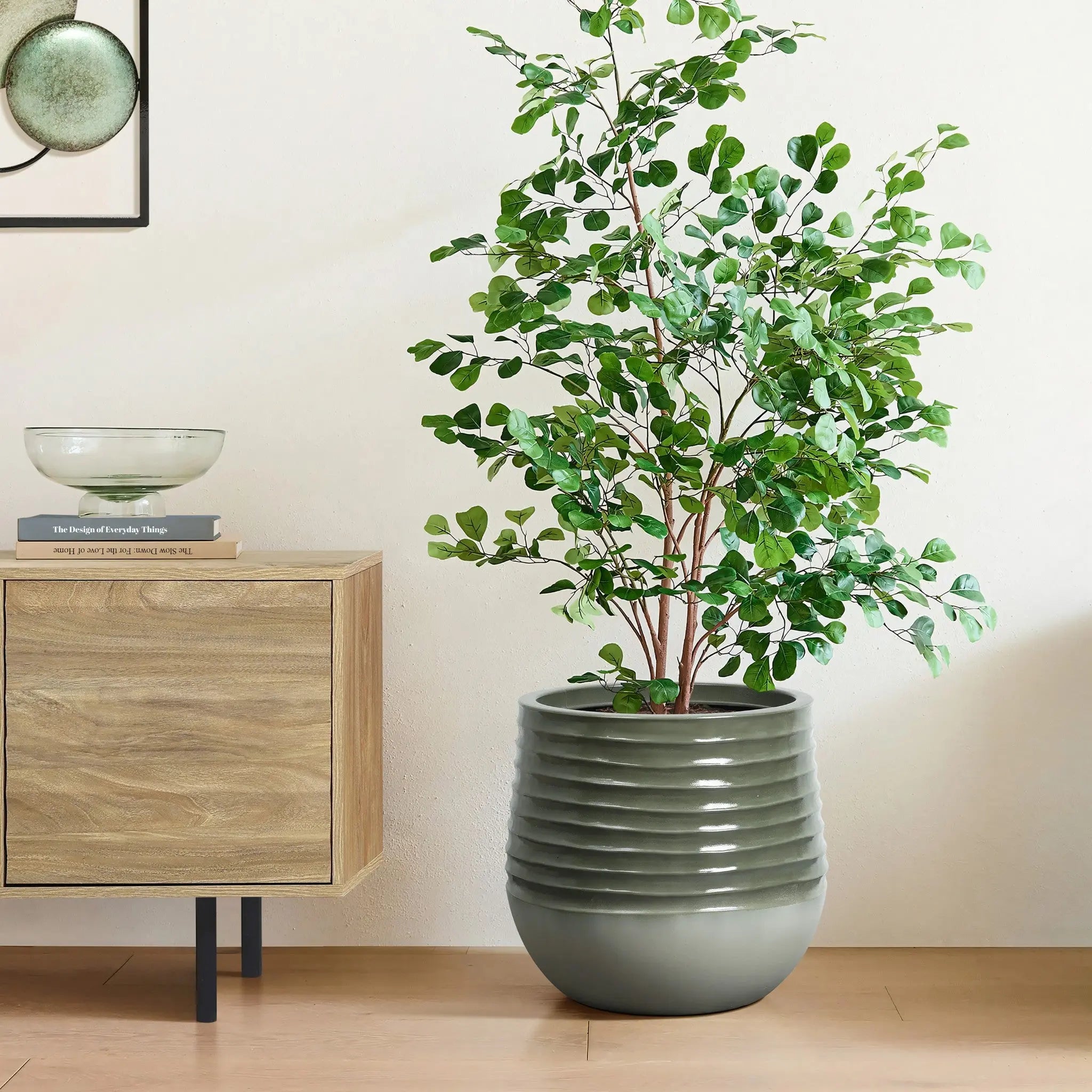 A leafy green plant luxuriates in a ribbed gray planter with a drainage hole beside a wooden cabinet on a light wood floor. The cabinet holds books and a glass bowl, seamlessly blending indoor and outdoor environments.