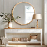 A round mirror with a scalloped design hangs above a wooden console table with three drawers. A lamp, vase with branches, and woven baskets decorate the table while natural light filters through a nearby window.
