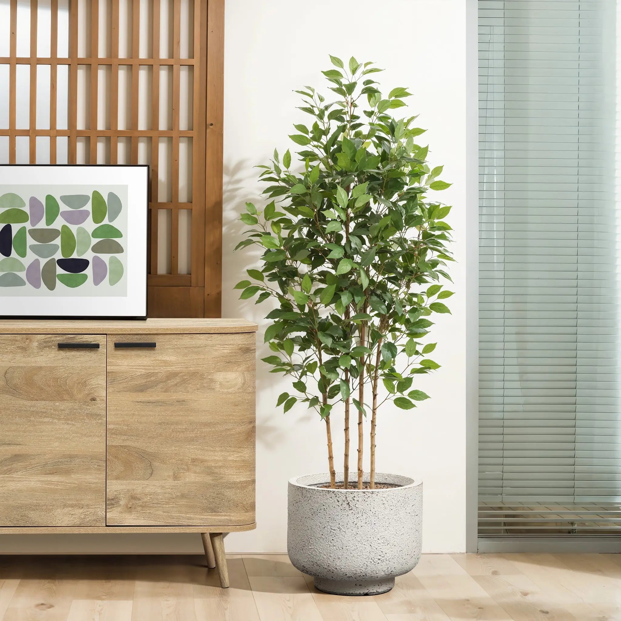 A tall, leafy potted plant in a white and gray planter stands on a wooden floor next to a wooden cabinet adorned with artwork. To the right, vertical blinds cover a glass panel, enhancing the retro minimalist decor of the room.