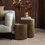 Two wooden teardrop accent tables add rustic charm beside a white textured sofa. The larger table holds stacked books and a decorative vase with dried leaves. The setting includes a patterned rug and wood-paneled wall, perfectly blending mid-century modern style with warmth.