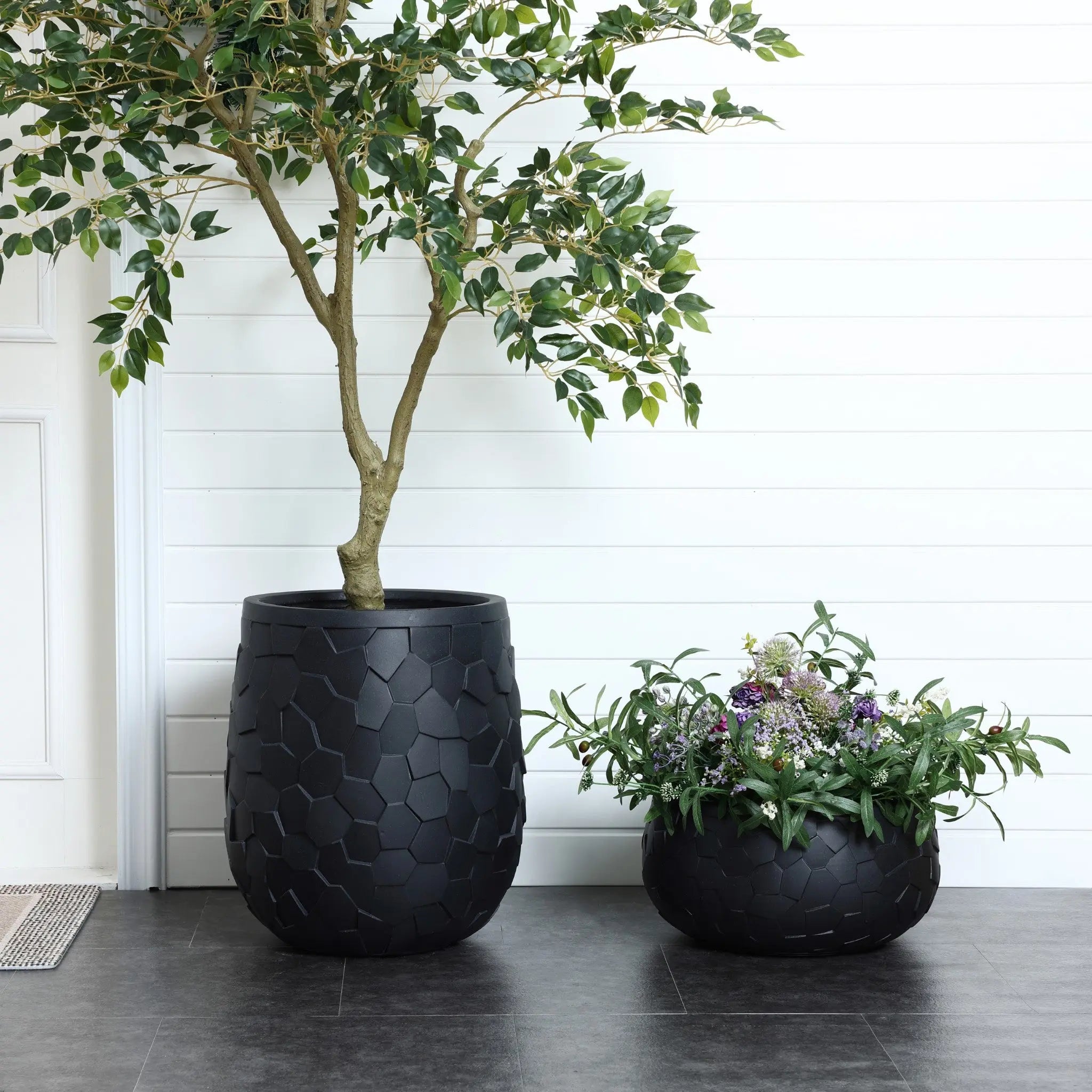 A tall potted tree in a LuxenHome black beehive pattern round tabletop planter, 17.5 wide, stands beside a shorter plant in a smaller matching pot. The scene is set against a white panel wall and door segment, with gray tiled flooring enhancing its durable charm.
