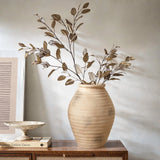A LuxenHome rustic beige ribbed terracotta urn vase, 13.2 high, with dried branches and leaves sits on a wooden table alongside a shallow ceramic dish and stacked books, enhancing the rustic decor against a plain wall.