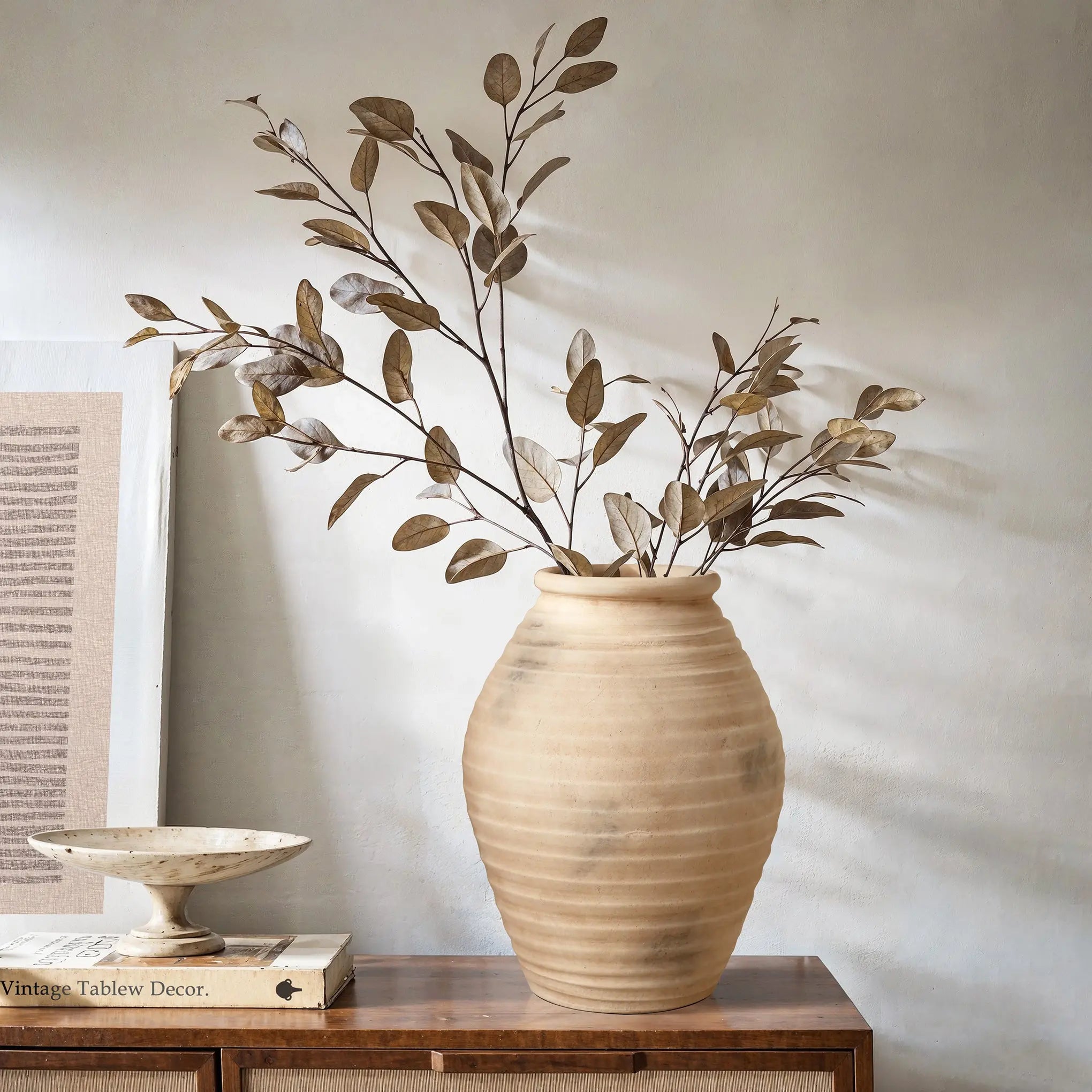 A LuxenHome rustic beige ribbed terracotta urn vase, 13.2 high, with dried branches and leaves sits on a wooden table alongside a shallow ceramic dish and stacked books, enhancing the rustic decor against a plain wall.
