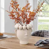The LuxenHome White ceramic bust head vase, 9.6 h, artfully designed with a sculpted head and fruit-like decor, holds autumn leaves on a wooden dining table set with plates and cutlery, offering a window view of greenery in the background.