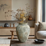 A LuxenHome rustic gold and brown terracotta urn vase (13.6 h) with dried branches adorns a wooden coffee table in a minimalist living room. A beige couch, neutral decor, pottery, and a large abstract painting complete the serene earth-toned setting.
