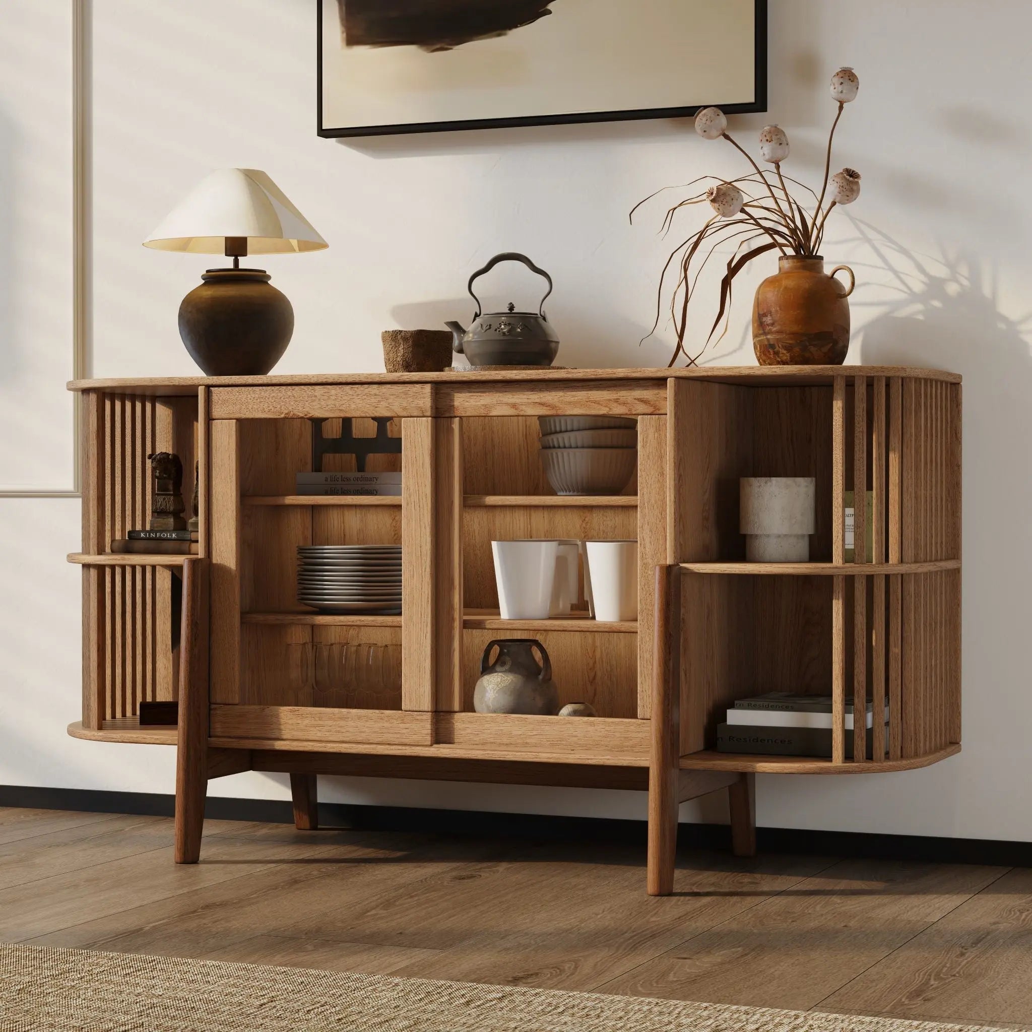 A cozy, well-lit room with wooden flooring features the LuxenHome Miko sideboard server cabinet with solid wood legs. Open shelves display pottery and dishes, while a table lamp and dried flowers in a vase sit on top. A framed abstract painting hangs above completing the mid-century modernist look.